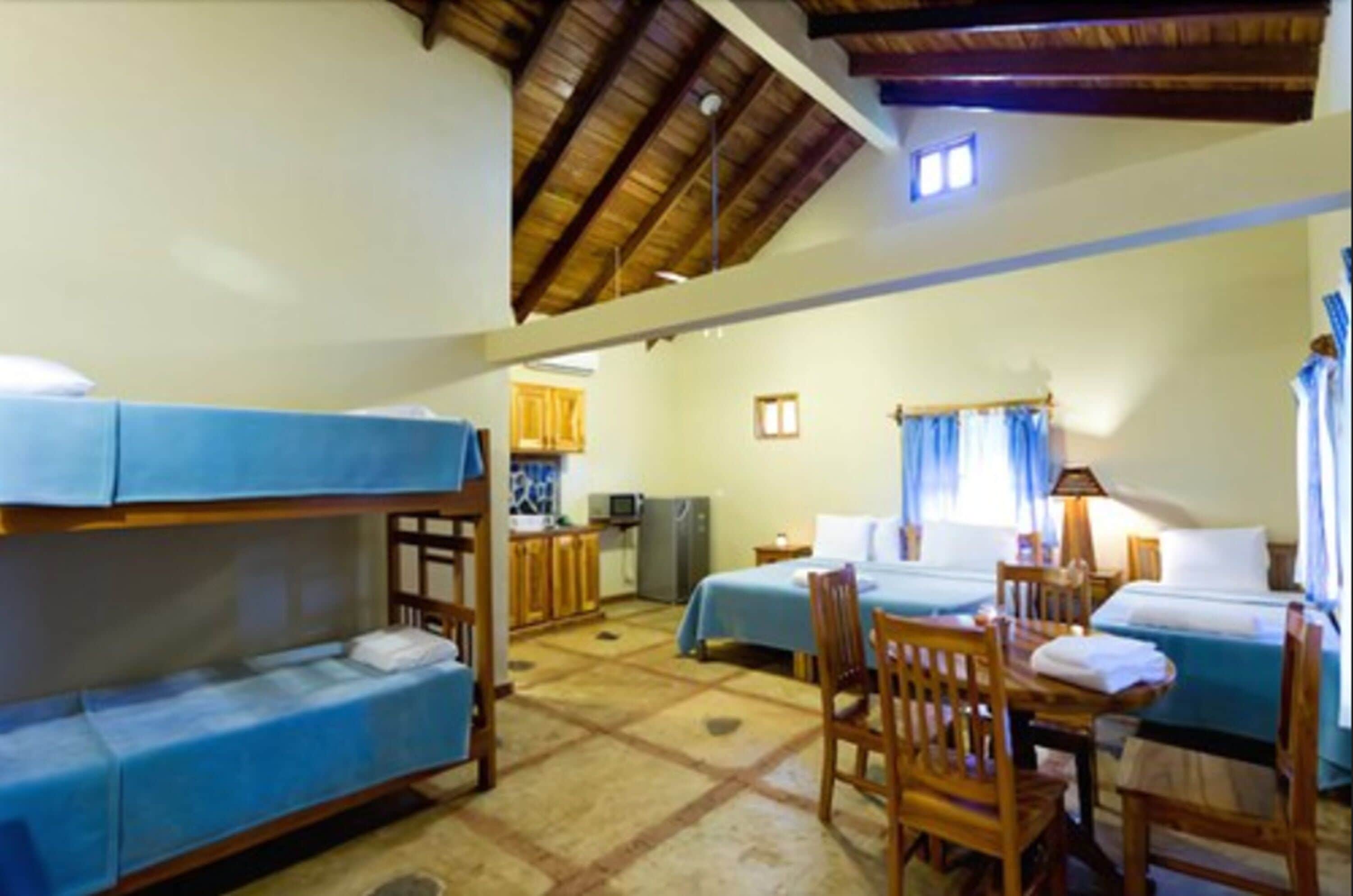 A room with bunk beds, a dining table with chairs, a kitchen area, and a window with curtains.