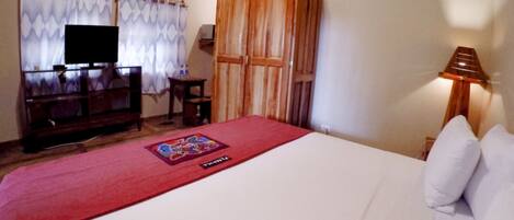 Habitación de hotel con una cama, un televisor sobre un mueble de madera, un armario de madera, una mesita pequeña y una lámpara.