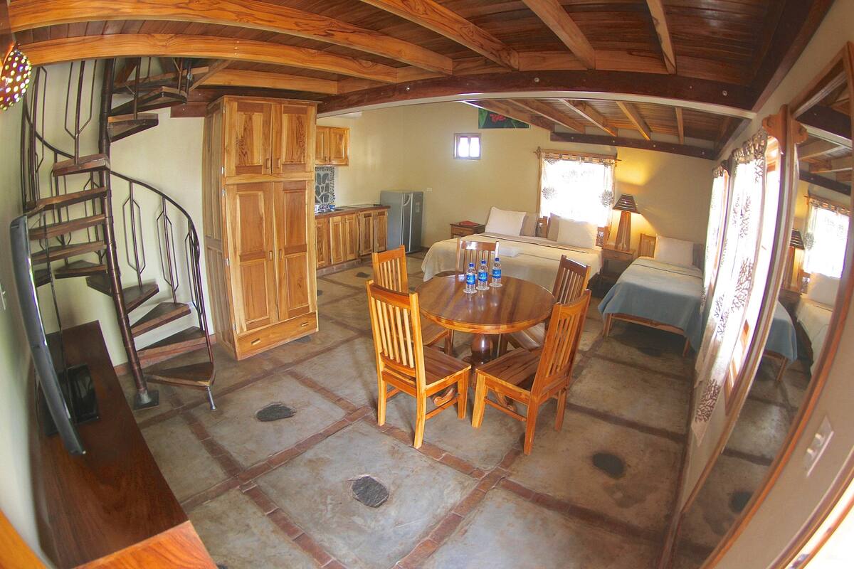 Een gezellige kamer met een houten eettafel, stoelen, een bed, een kleine keuken en een spiltrap.