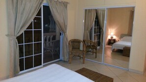 Une chambre dotée d’une grande porte-fenêtre coulissante donnant sur un balcon, avec un lit, une chaise et une table de chevet avec une lampe.