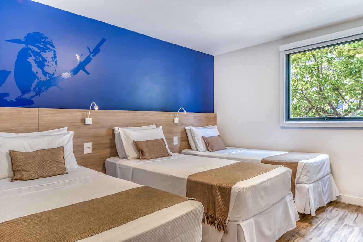 A hotel room with three beds, a blue mural on the wall, and a window with a view of trees.