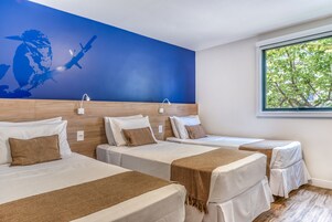 Habitación de hotel con tres camas, un mural azul en la pared y una ventana con vistas a árboles.