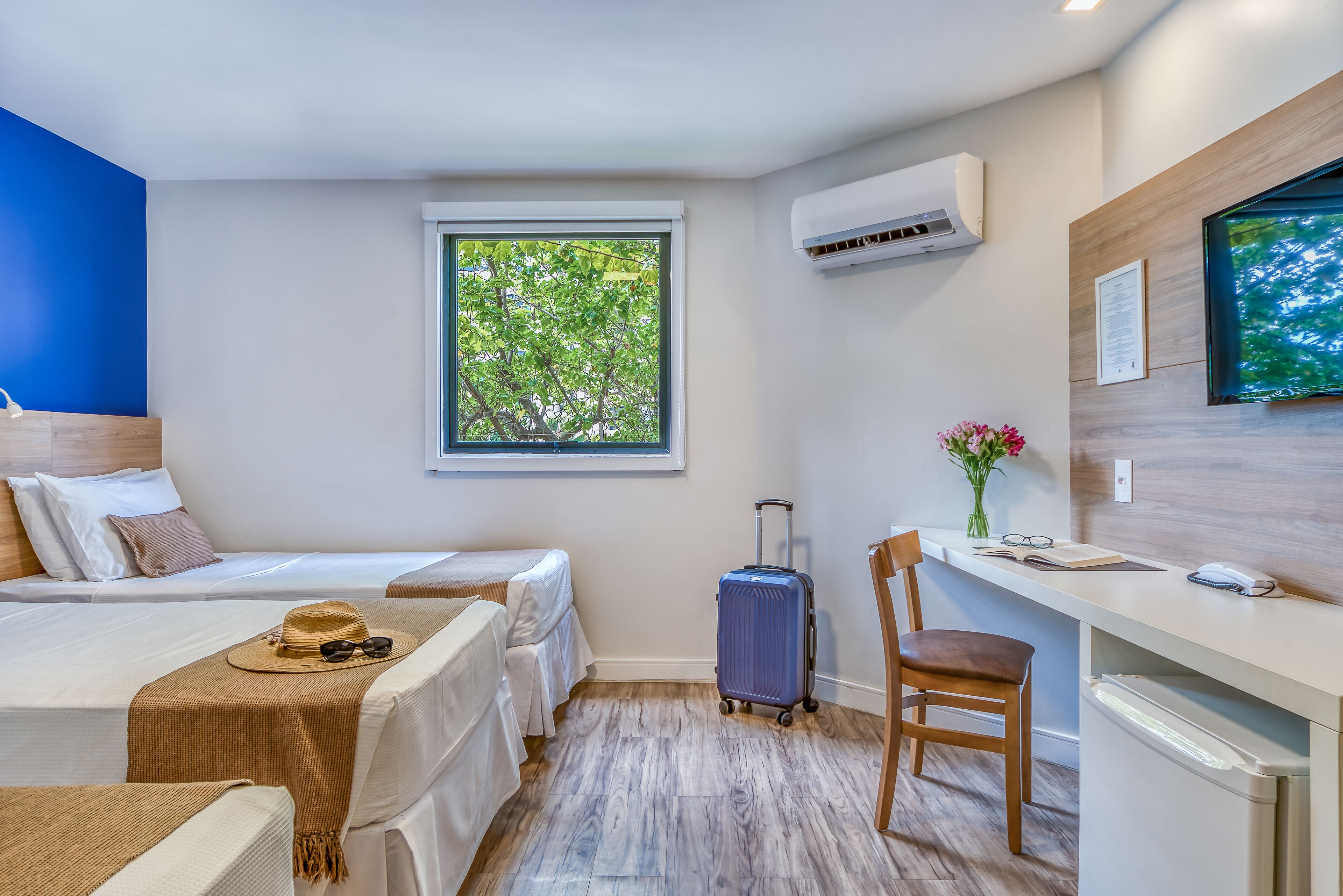 Une chambre d’hôtel avec deux lits, un bureau, une chaise, une valise et une fenêtre donnant sur des arbres.