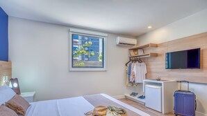 A hotel room with a bed, a desk, a TV, a suitcase, and a window with a view of buildings and trees. - Cais da Praia Hotel (Maceió)