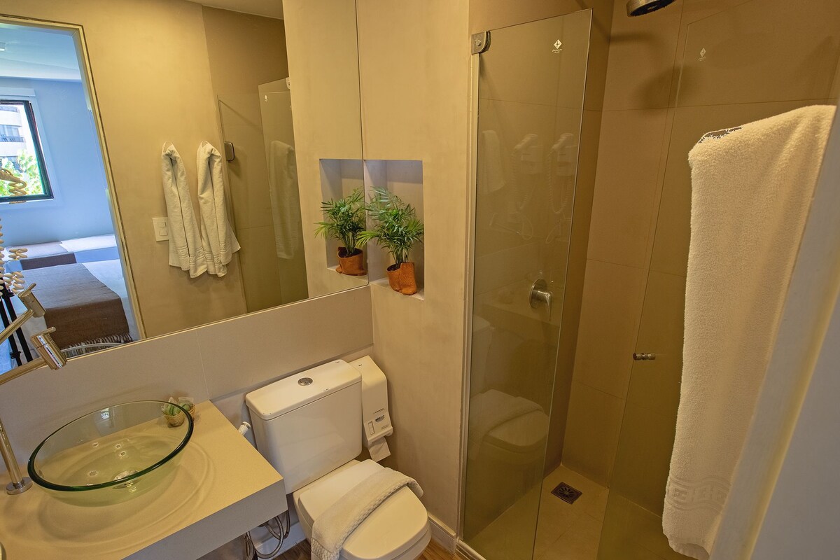 A modern bathroom with a glass sink, toilet, and a glass-enclosed shower. There are two potted plants on the wall and a towel hanging on the door.