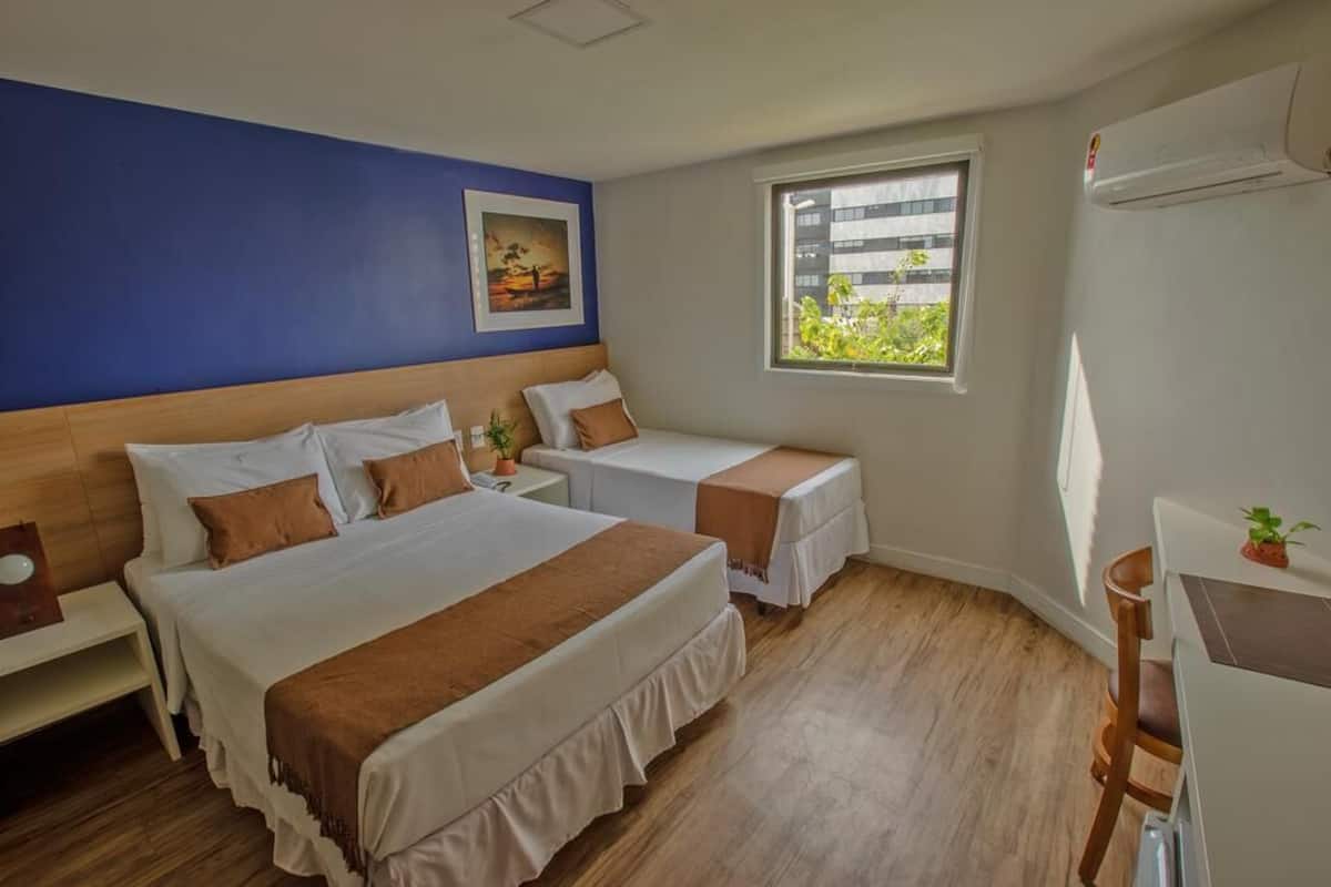 A hotel room with two beds, a desk, a chair, a window with a view of buildings, and an air conditioning unit.