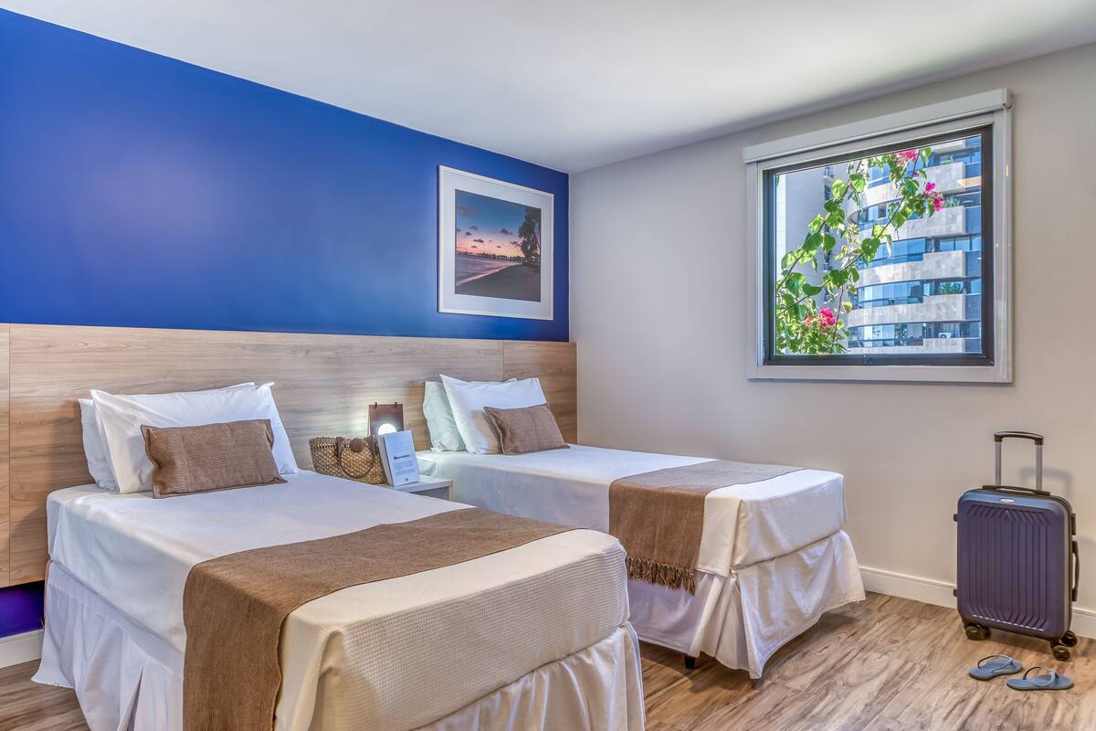 A hotel room with two beds, a wooden headboard, a nightstand, a framed picture on the wall, a window with a view of buildings and flowers, and a suitcase on the floor.