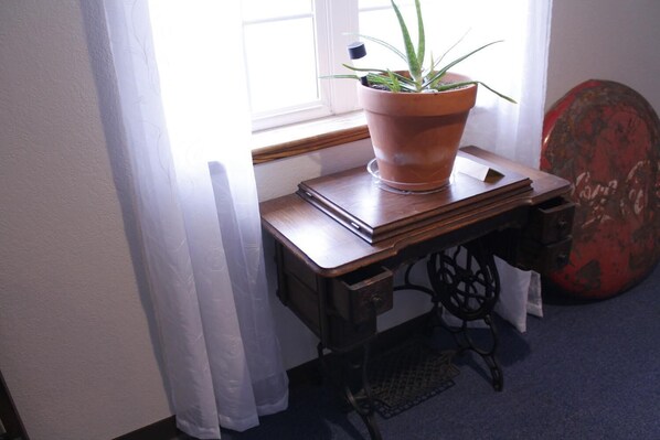 Una ventana con cortinas translúcidas, una planta en maceta, una pila de libros y una máquina de coser antigua sobre una mesa de madera.