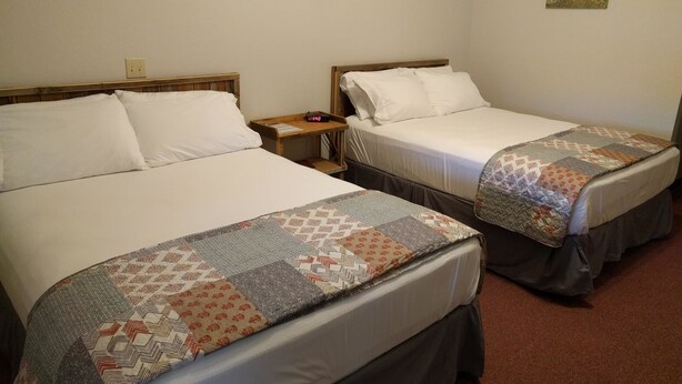 Two single beds with white linens and a quilt, a bedside table with a clock, and a wall-mounted light fixture.