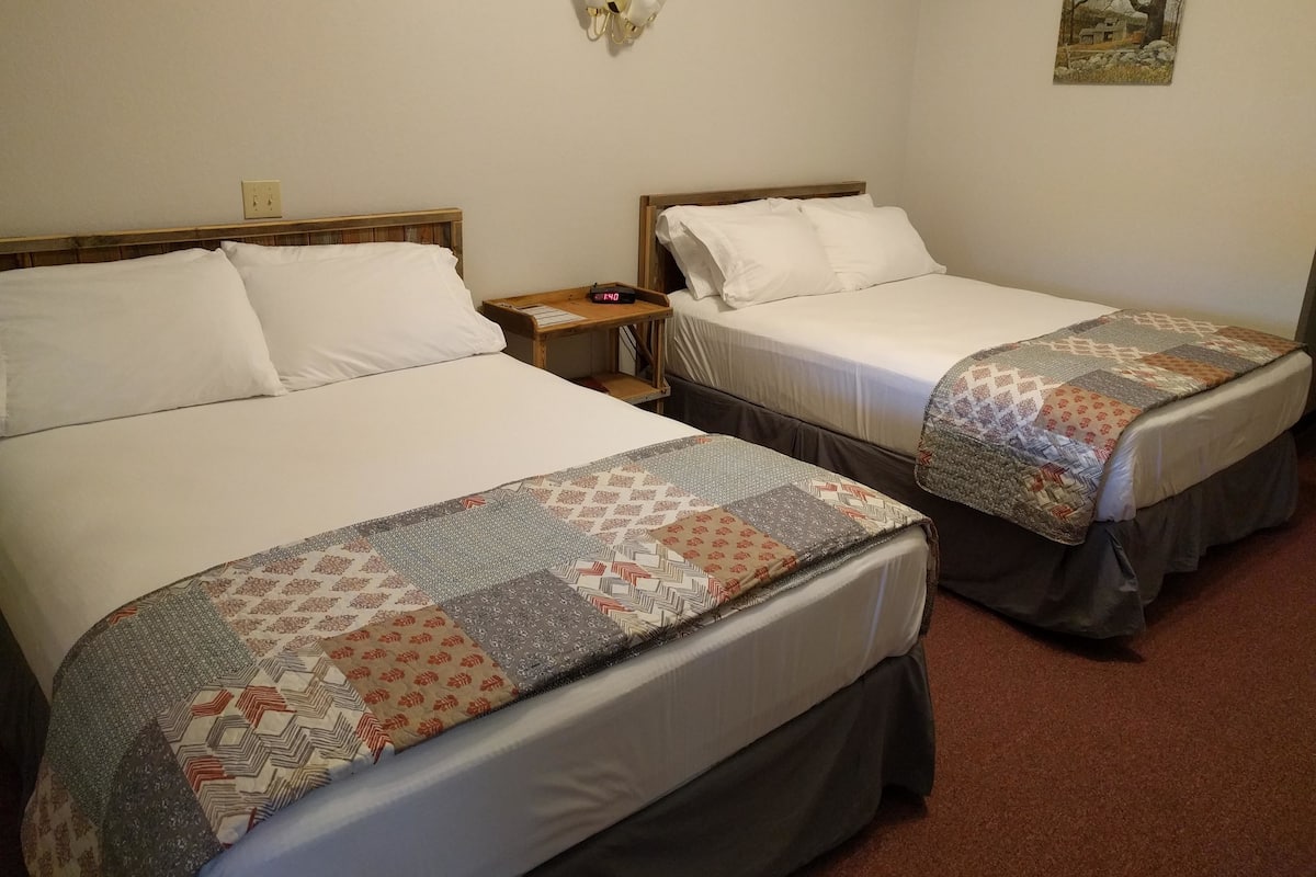 Two single beds with white linens and a quilt, a bedside table with a clock, and a wall-mounted light fixture.
