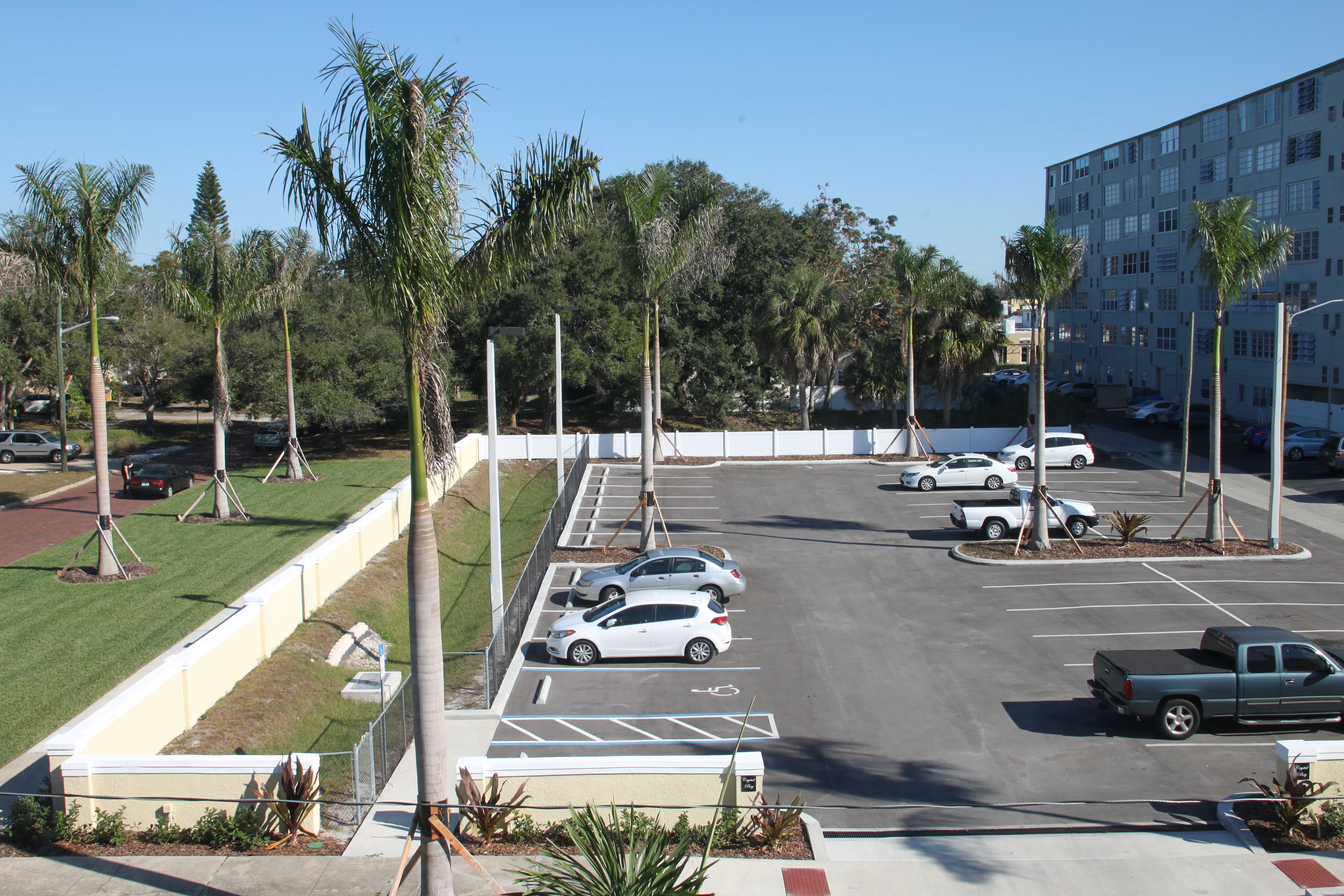 A parking lot with several cars and a truck, surrounded by palm trees and a building in the background.