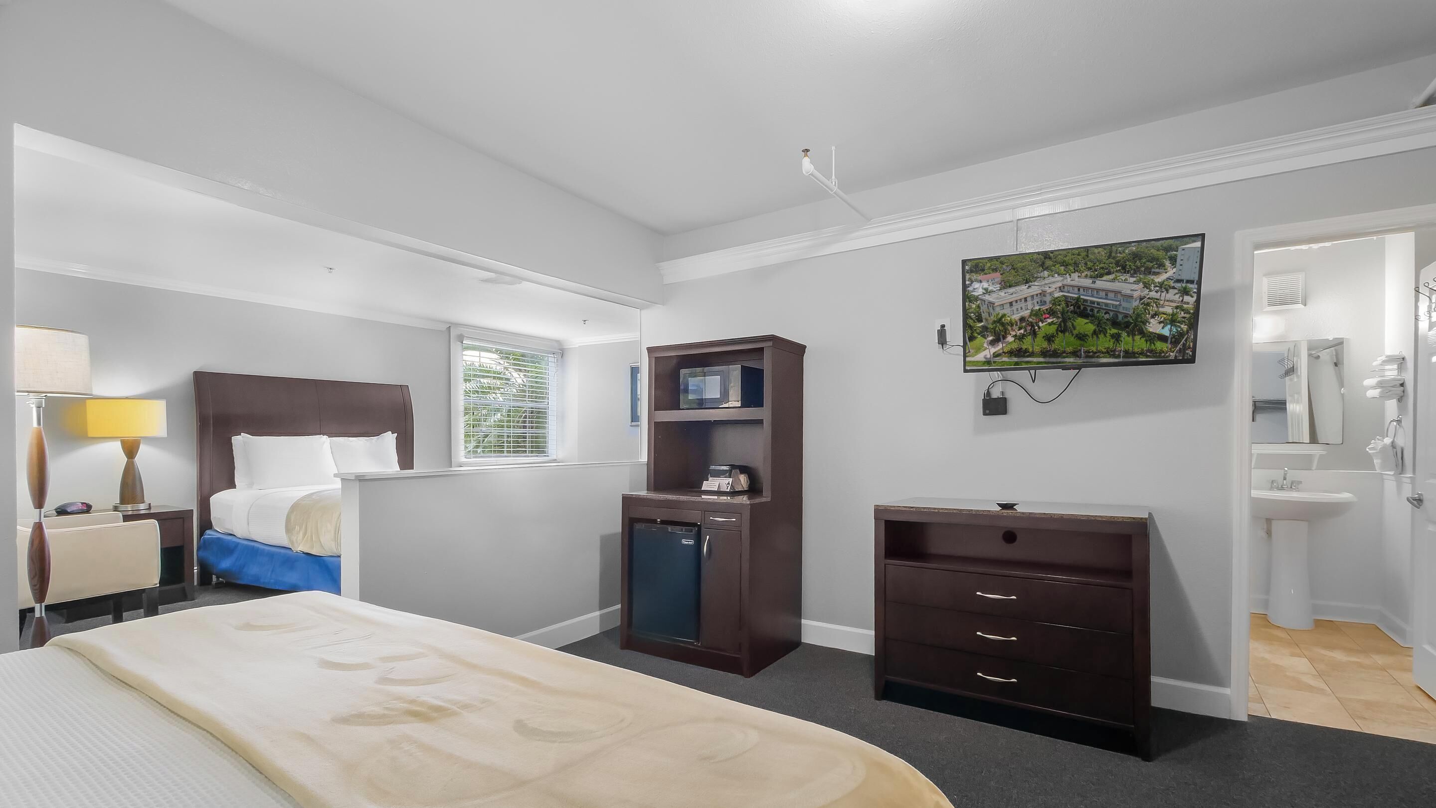 A hotel room with a bed, bedside table with a lamp, a dresser, a desk with a computer, a window with blinds, a painting on the wall, and a bathroom with a sink and mirror.