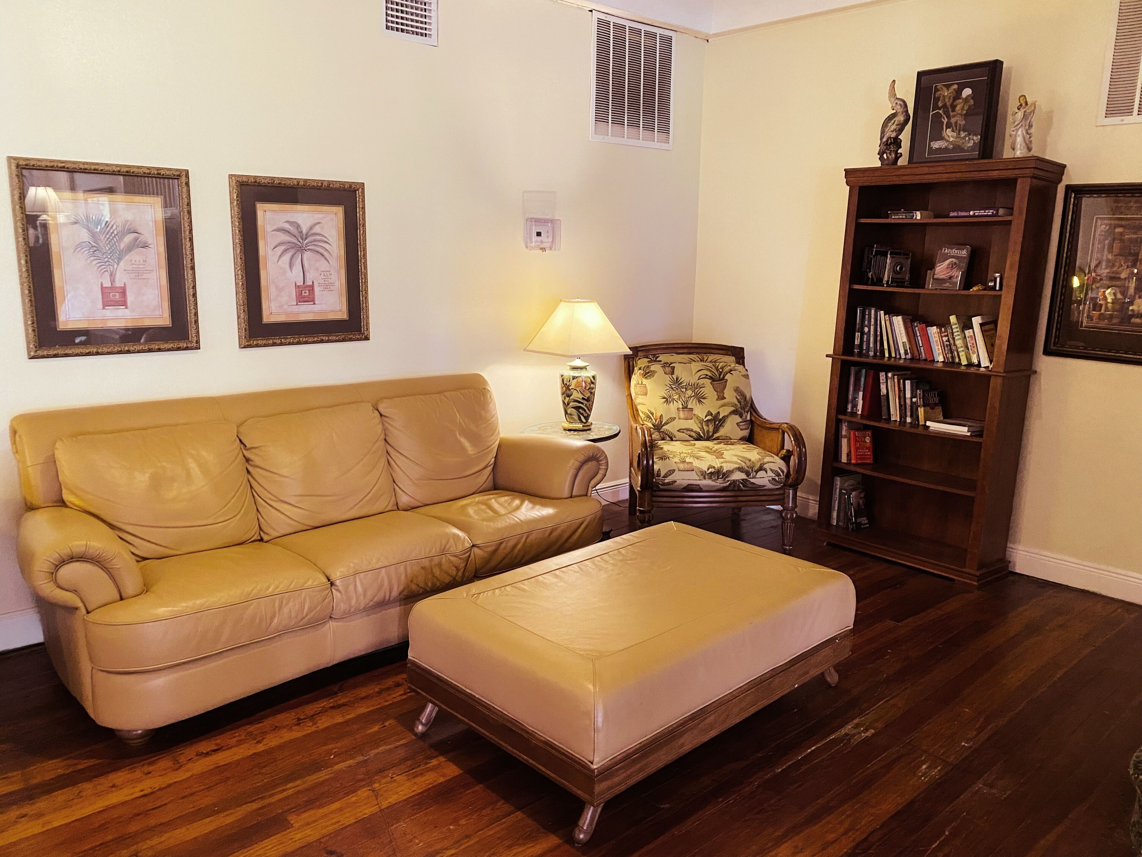 A living room with a beige sofa, a matching ottoman, a patterned armchair, a side table with a lamp, a bookshelf, and framed pictures on the wall.