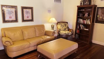 A living room with a beige sofa, a matching ottoman, a patterned armchair, a side table with a lamp, a bookshelf, and framed pictures on the wall.