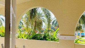 Une chaise blanche se trouve dans un espace extérieur abrité, doté de fenêtres en arc qui donnent sur un jardin où poussent différentes plantes et arbres.