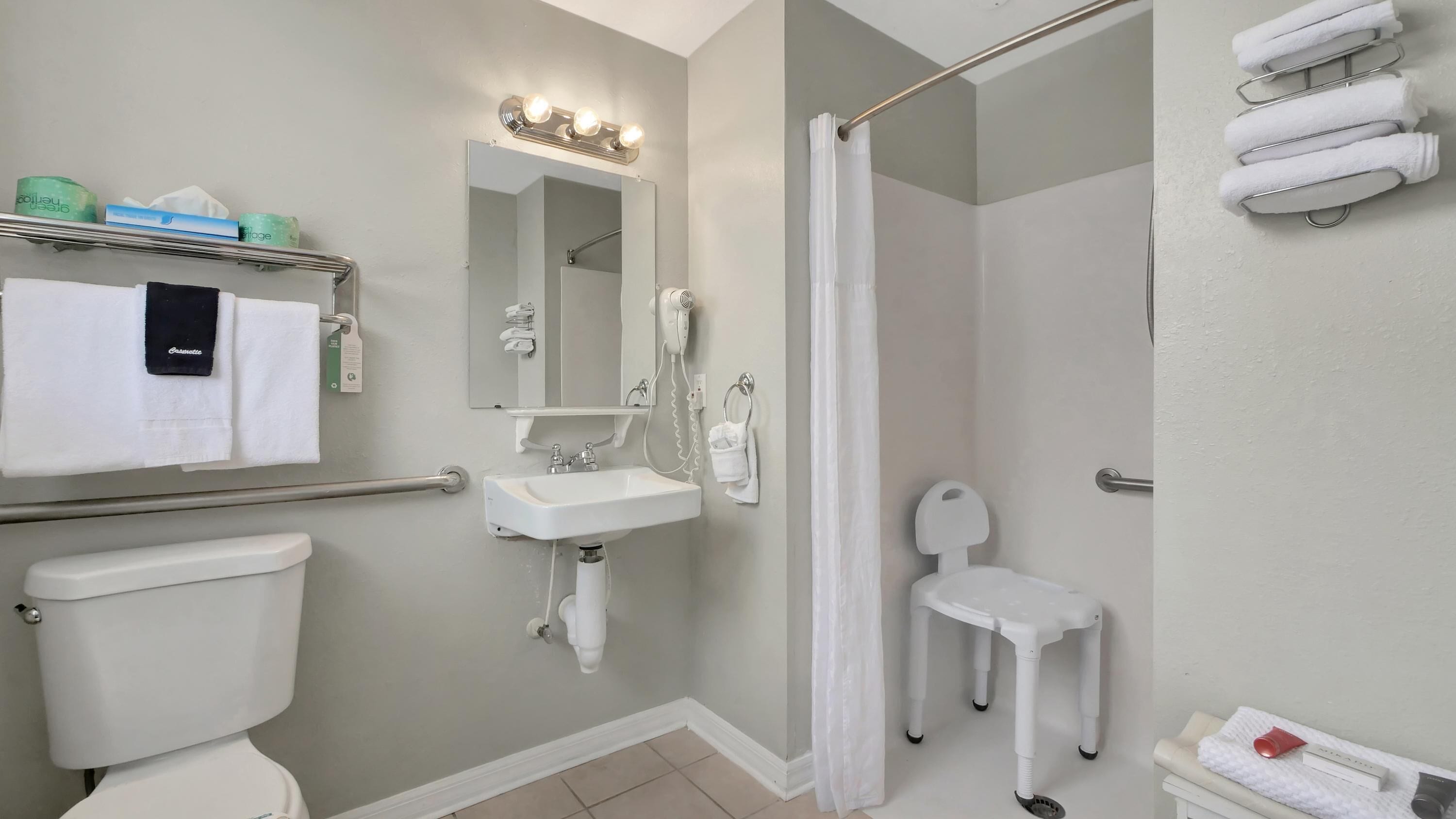 A bathroom with a toilet, sink, shower, and a small white chair.