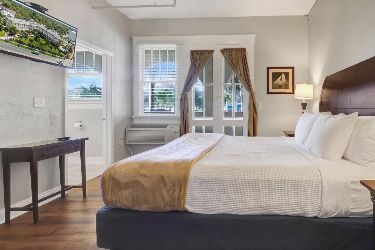 Une chambre d’hôtel bien rangée, avec un lit, une table de chevet, une lampe et une vue sur un immeuble par la fenêtre.