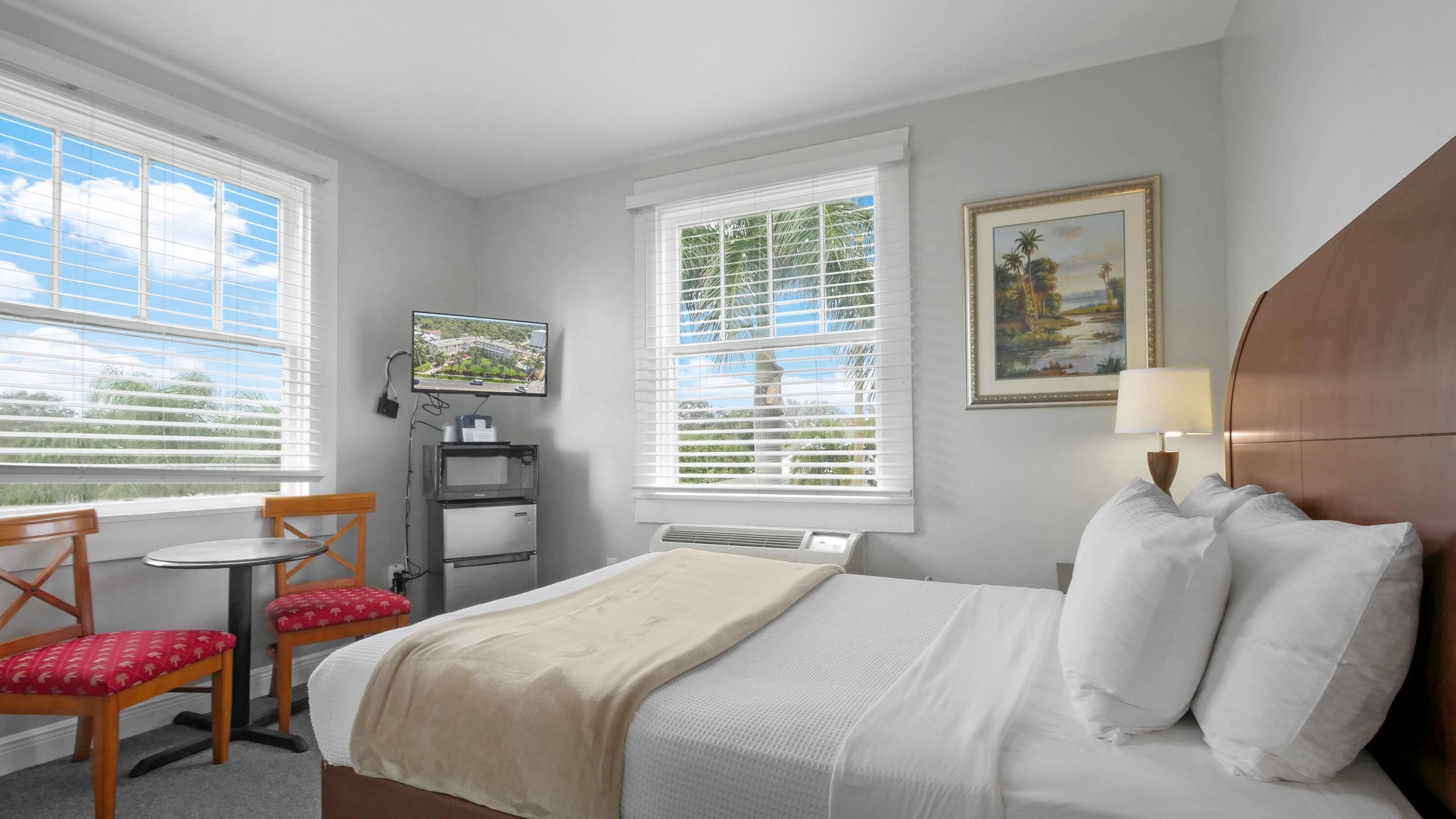 A hotel room with a bed, a desk with a chair, a television, and two windows with blinds.