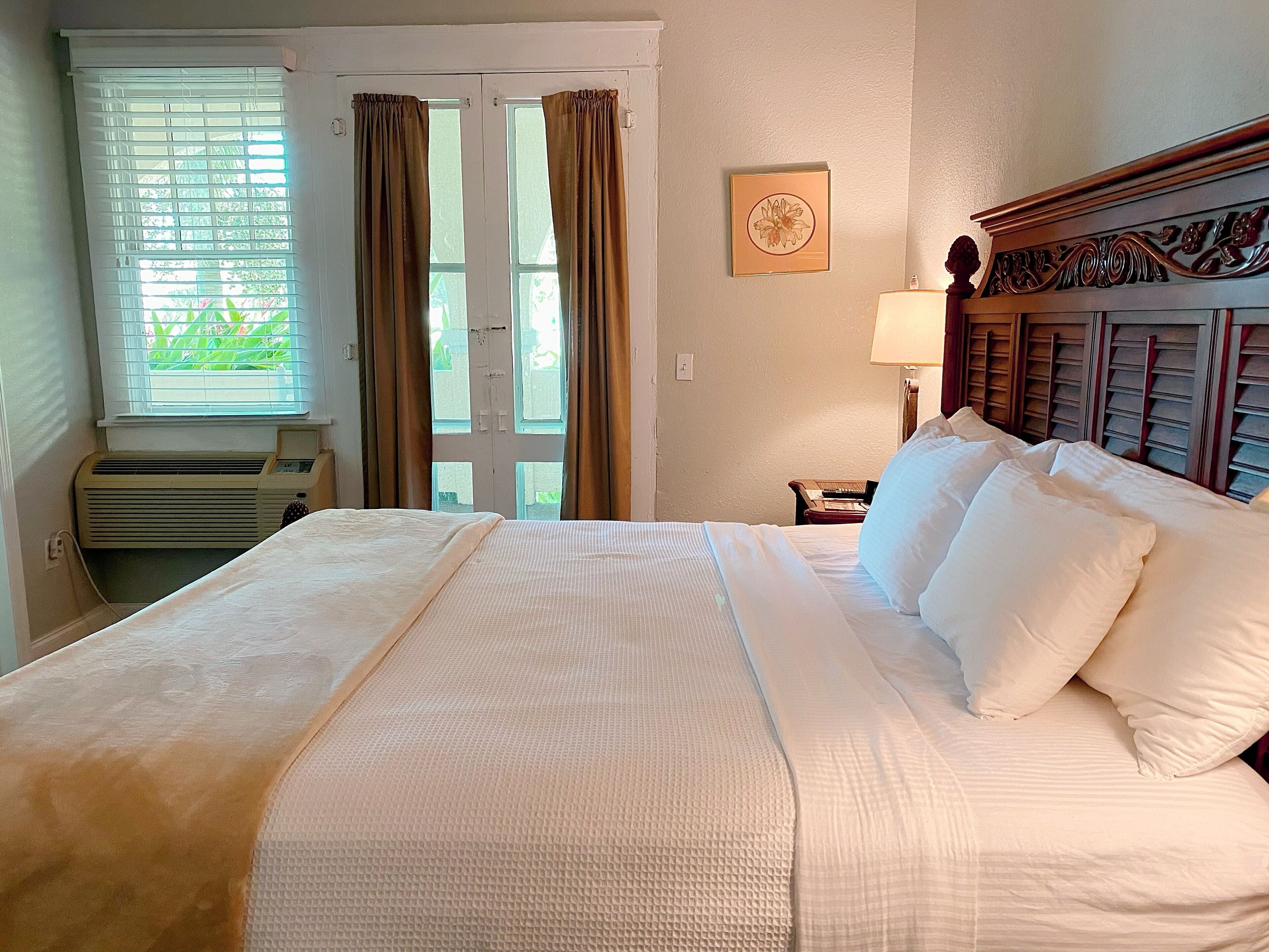 A neatly made bed with white linens, a window with blinds, a wall-mounted air conditioner, and a framed picture on the wall.