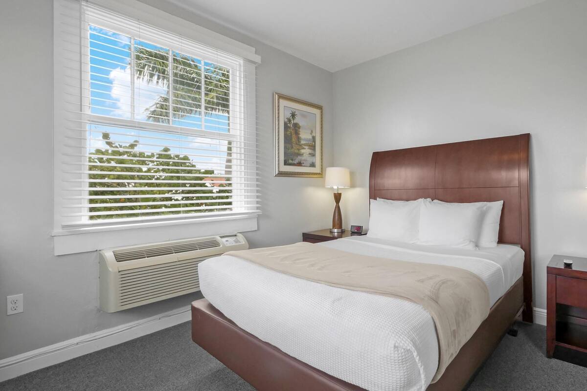 Une chambre d’hôtel bien aménagée, avec un grand lit, deux tables de chevet avec des lampes, une fenêtre avec des stores, un climatiseur fixé au mur et un tableau encadré.