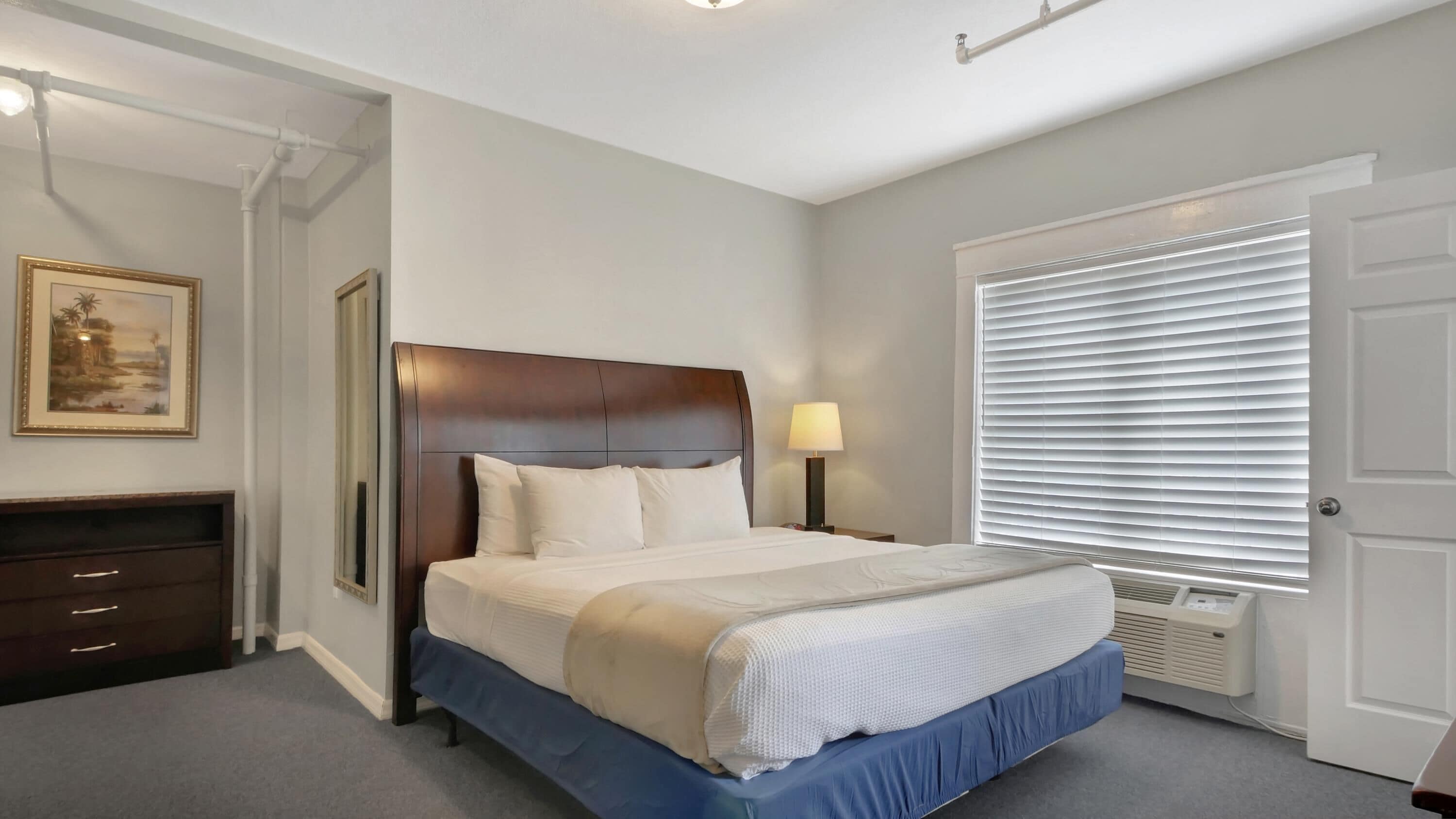 A neatly arranged bedroom with a large bed, a bedside table with a lamp, a dresser, and a window with blinds.