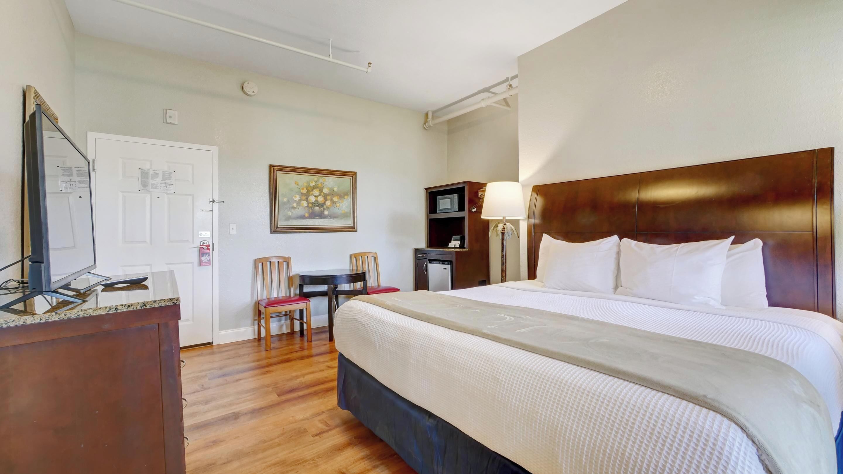 Quarto de hotel com cama grande, escrivaninha com computador, cadeira, mesinha, micro-ondas e um quadro na parede.