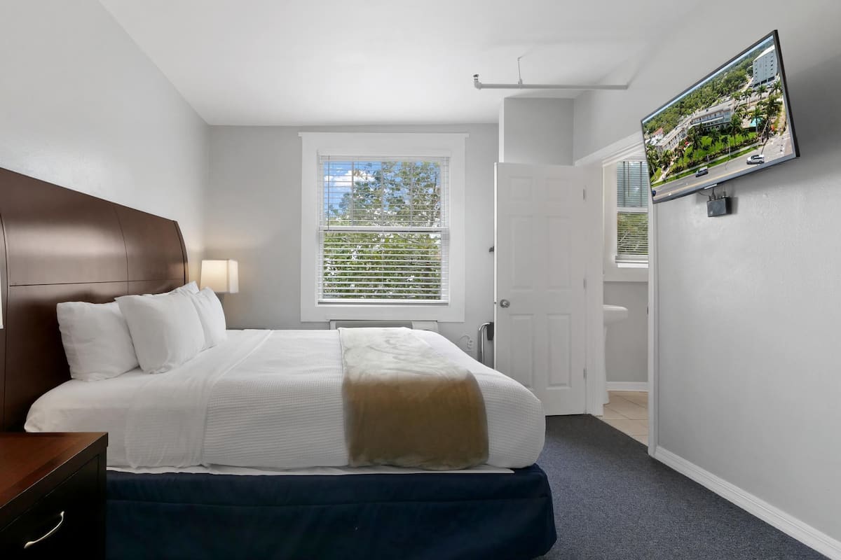 Une chambre à coucher comprenant un lit, une table de chevet, un téléviseur fixé au mur et une fenêtre avec des stores.