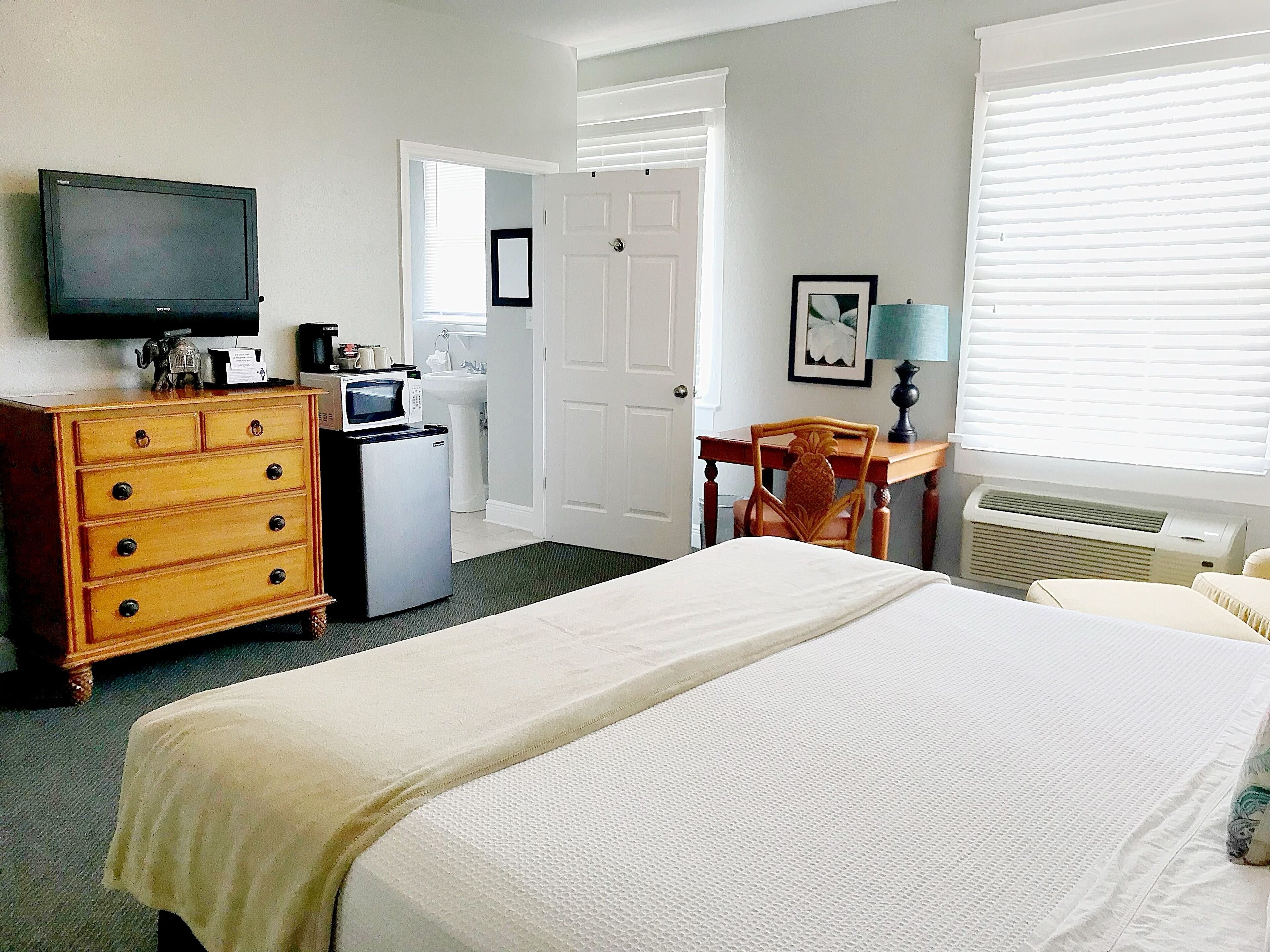 A hotel room with a bed, a dresser, a television, a microwave, a chair, a desk, and a window with blinds.
