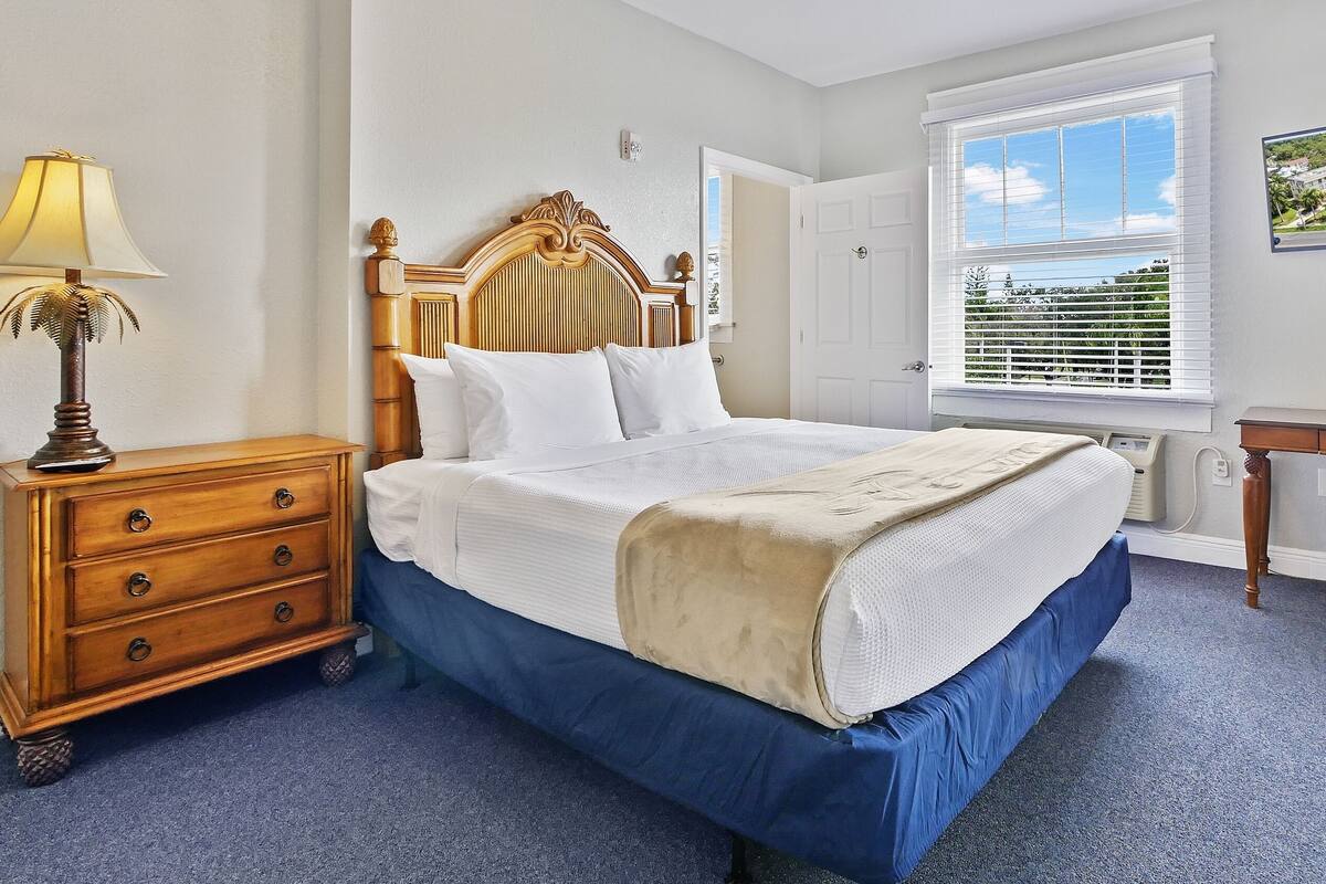 Une chambre à coucher comprenant un lit, une commode, une chaise, une fenêtre avec des stores et un téléviseur fixé au mur.