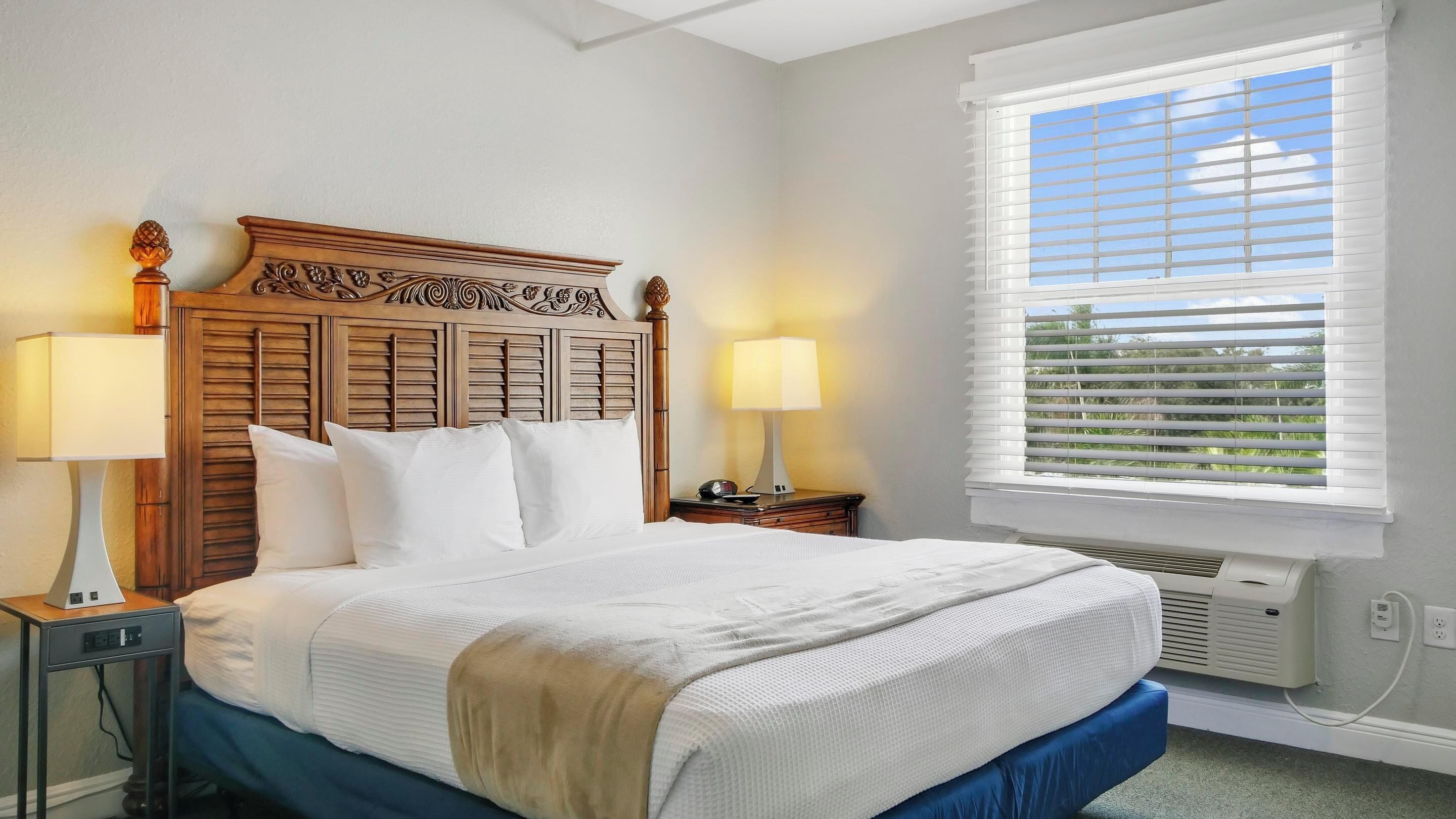 Una cama bien hecha con cabecera de madera, dos lámparas de noche y una ventana con persianas.