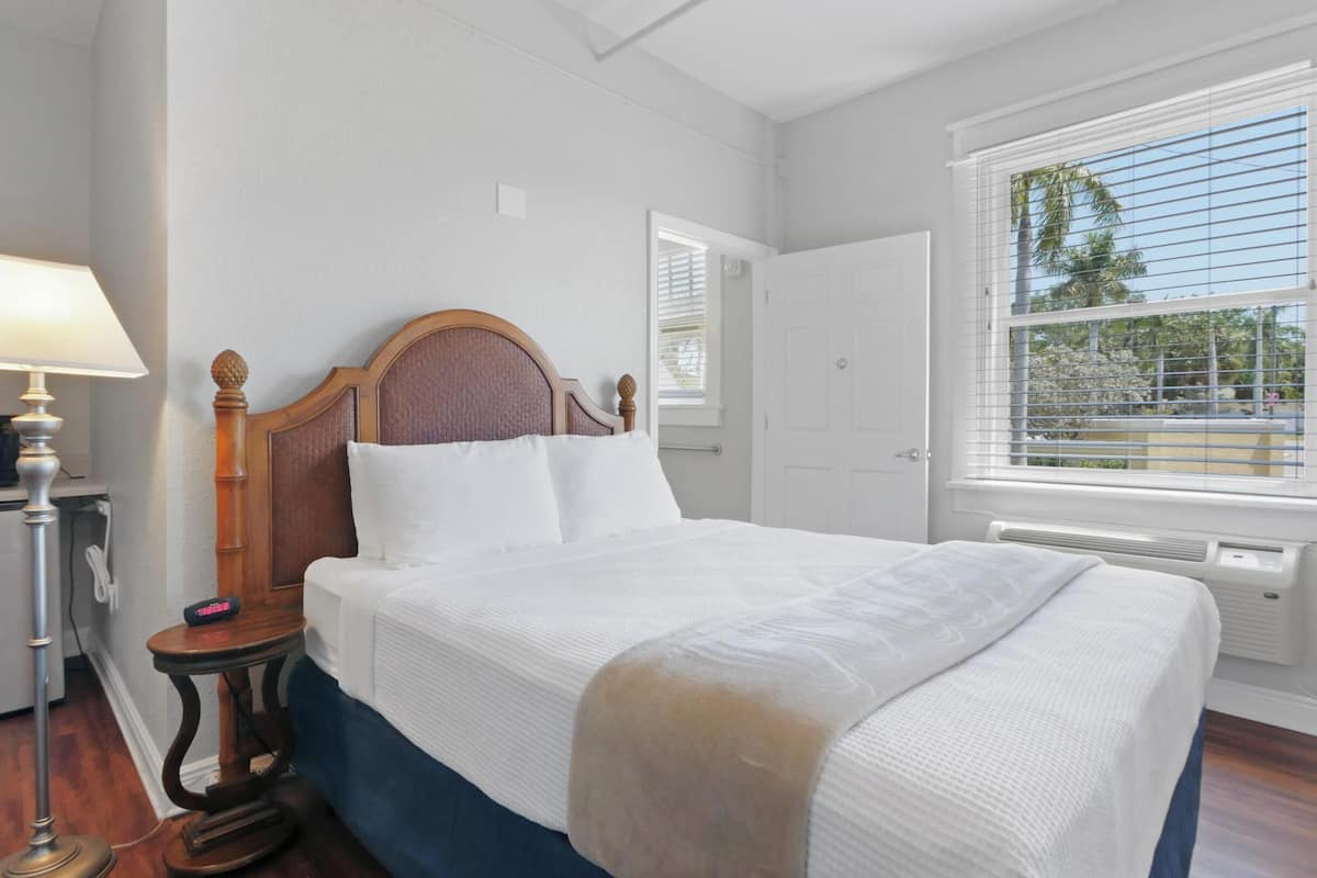 Une chambre à coucher comprenant un lit, une table de chevet, une lampe, une fenêtre avec des stores et une porte.