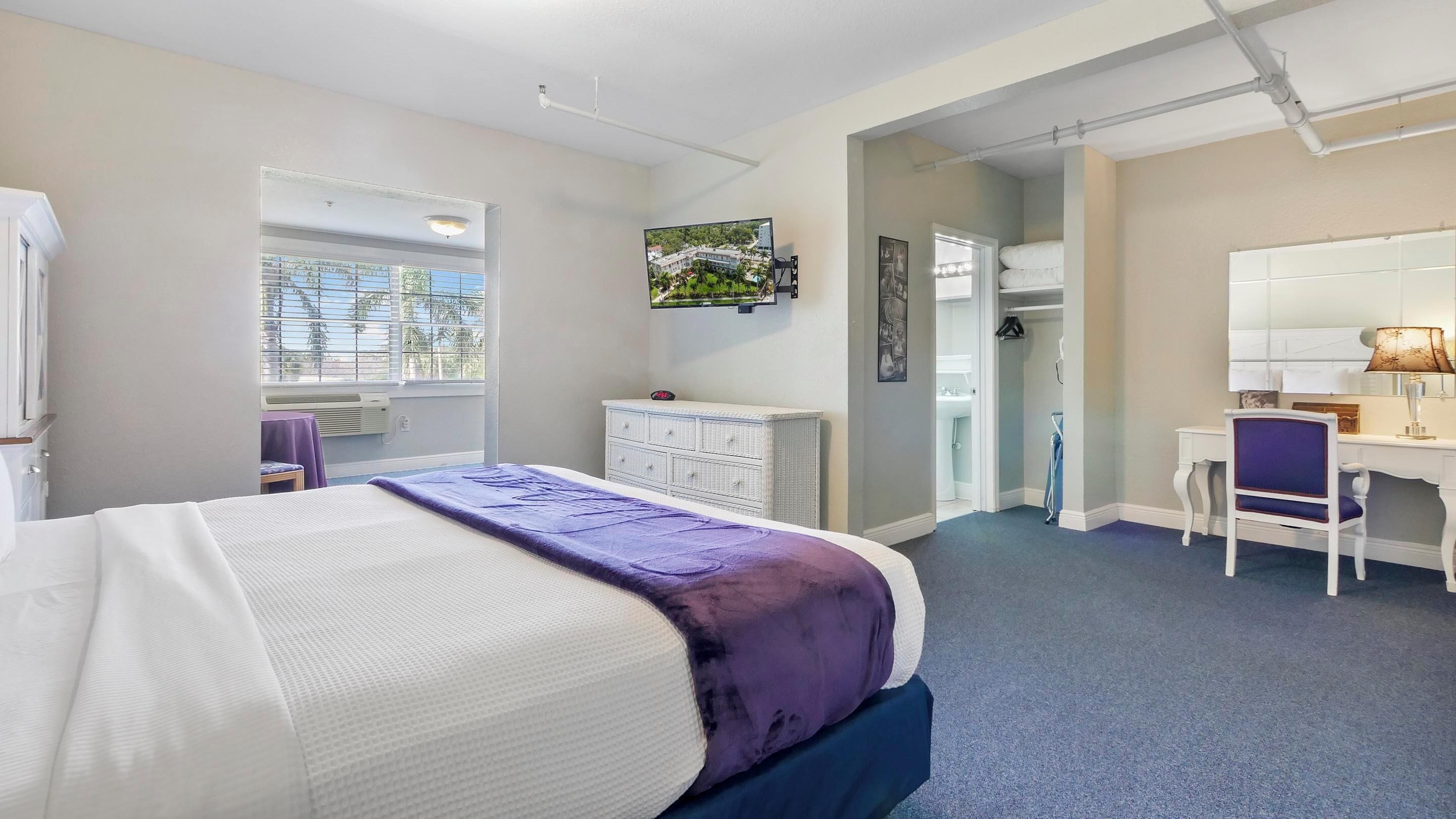 A hotel room with a bed, a desk, a chair, a dresser, a window, and a TV.