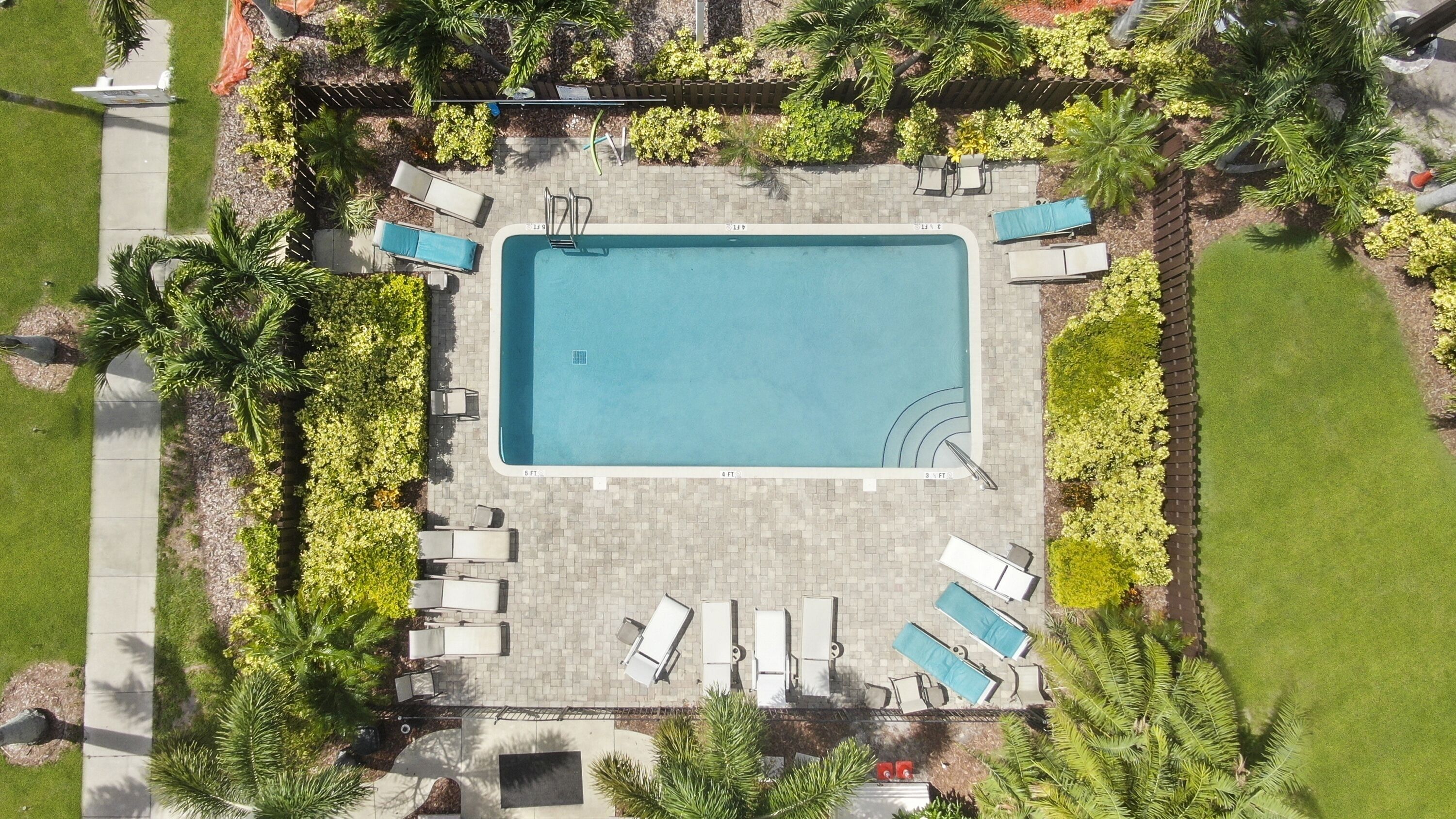 An aerial view of a rectangular swimming pool surrounded by a paved deck, with lounge chairs and a small garden area.