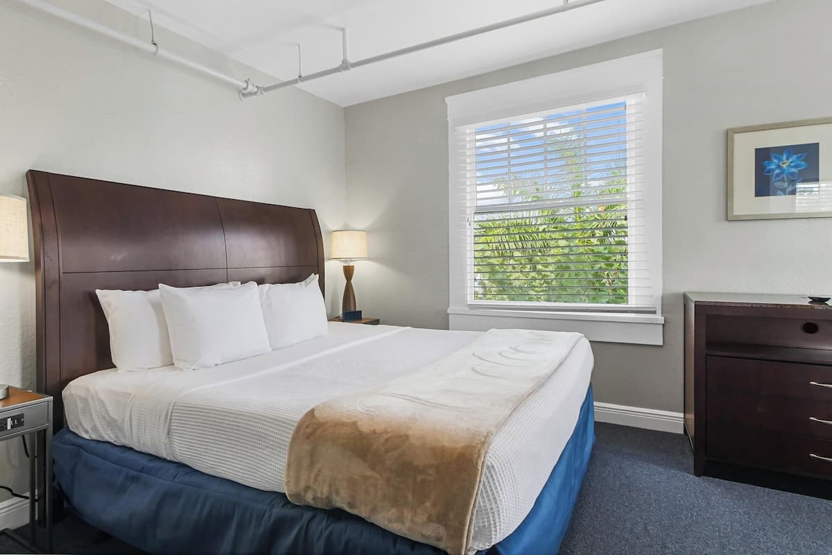 Une chambre d’hôtel avec un grand lit, deux tables de chevet avec des lampes, une commode et une fenêtre avec des stores.
