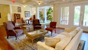 Un hall d’entrée spacieux, avec un grand canapé blanc, plusieurs fauteuils, une table basse et un tapis à motifs. Plusieurs portes donnent sur l’extérieur, et on peut apercevoir une plante en pot.