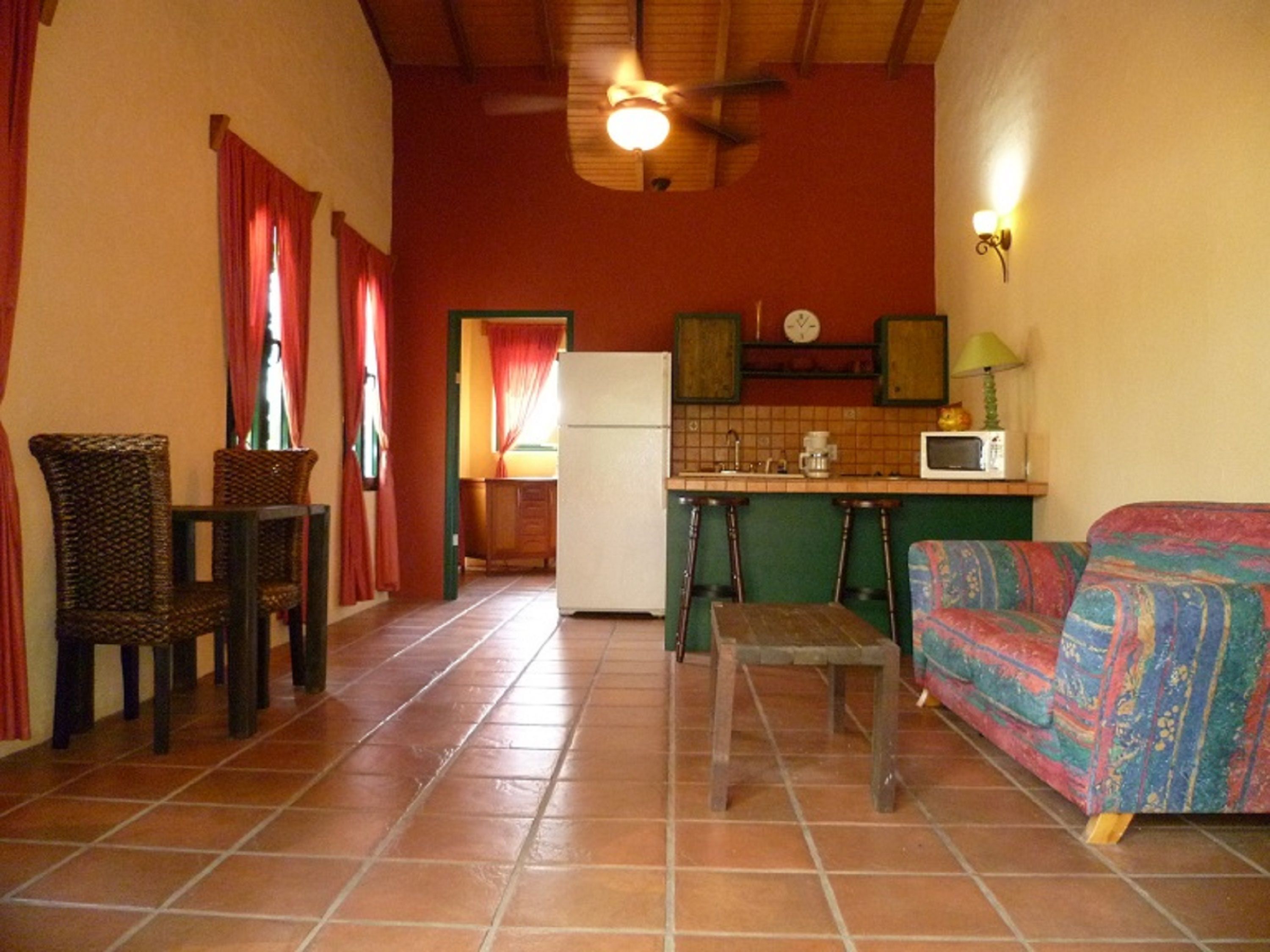 A compact living space with a kitchen, a dining area with wicker chairs, a colorful sofa, and a small table.