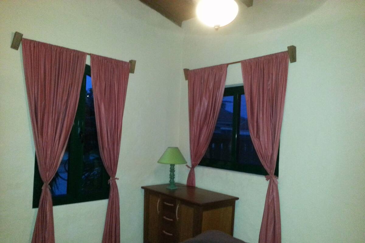Une pièce avec deux fenêtres, chacune ornée de rideaux roses maintenus par des liens, une petite table de chevet en bois sur laquelle se trouve une lampe verte, et un lit recouvert d’une couverture sombre.