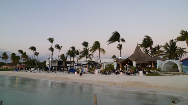 A beachfront resort with palm trees, beachside seating, and thatched-roof structures.