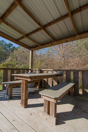 Una terraza de madera cubierta con mesas de picnic y bancos, rodeada de árboles.