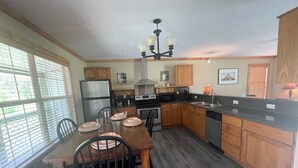 A modern kitchen with wooden cabinets, a dining table with chairs, a refrigerator, a stove, and a chandelier. - The Stagecoach Inn of Woodville (Woodville)