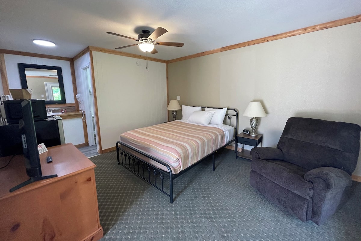 A hotel room with a bed, a desk, a chair, a ceiling fan, and a small table with a lamp.