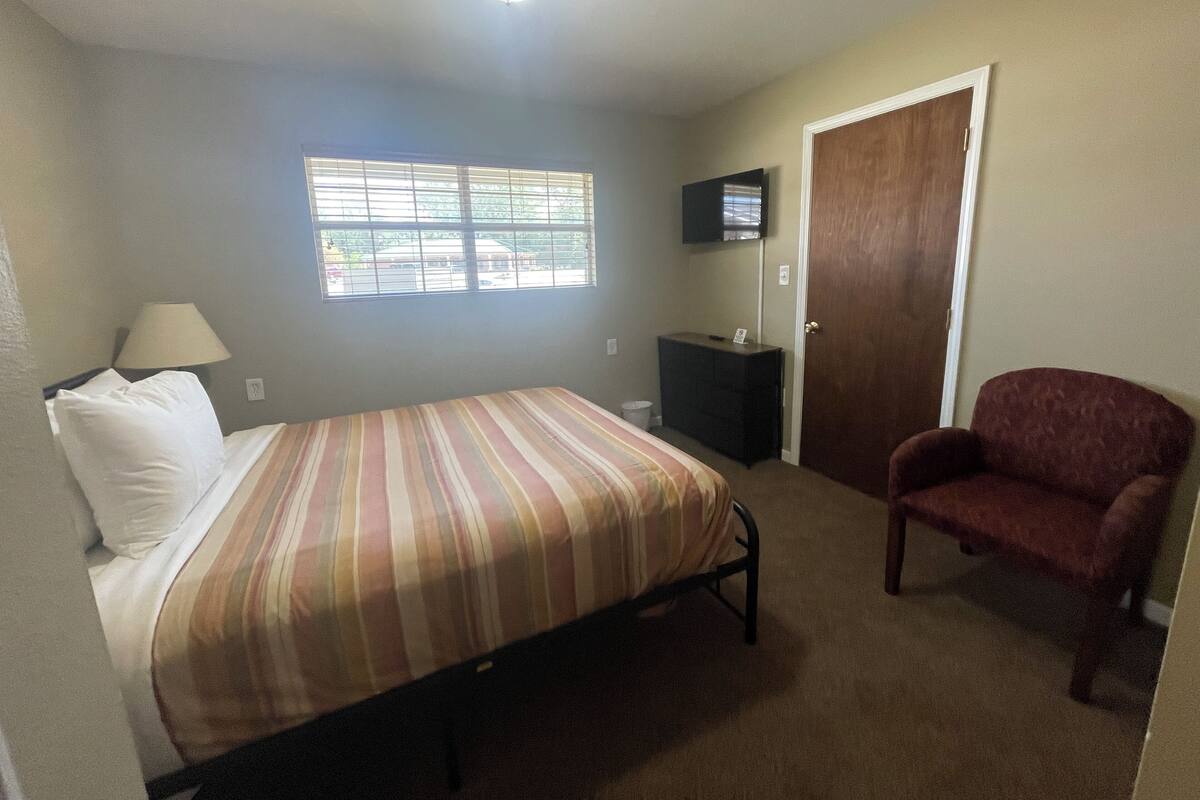 A bedroom with a bed, a chair, a desk, a TV, and a window.