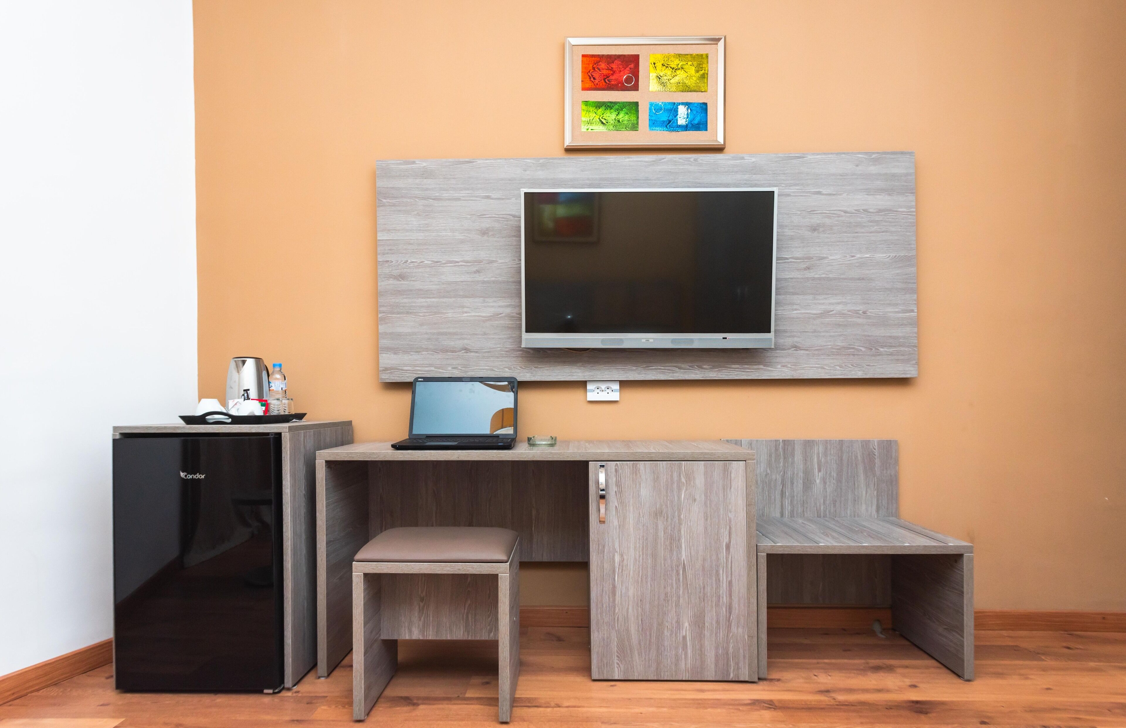 A compact room with a built-in TV, a small fridge, a desk, and a bench.
