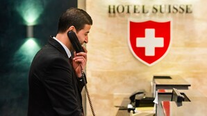A man in a suit talking on a telephone at a hotel reception desk with a sign reading "HOTEL SUISSE" and a red cross emblem. - Hôtel Suisse (Algiers)
