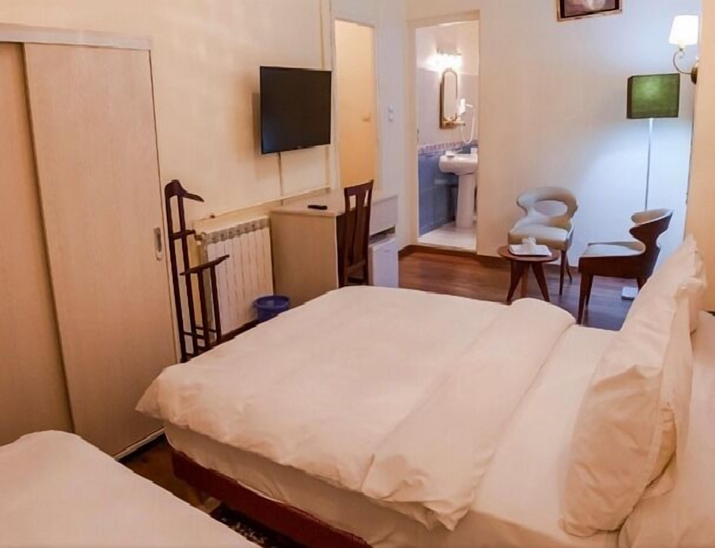 Una habitación de hotel con cama, televisor montado en la pared, baño con lavamanos y espejo, y una silla.
