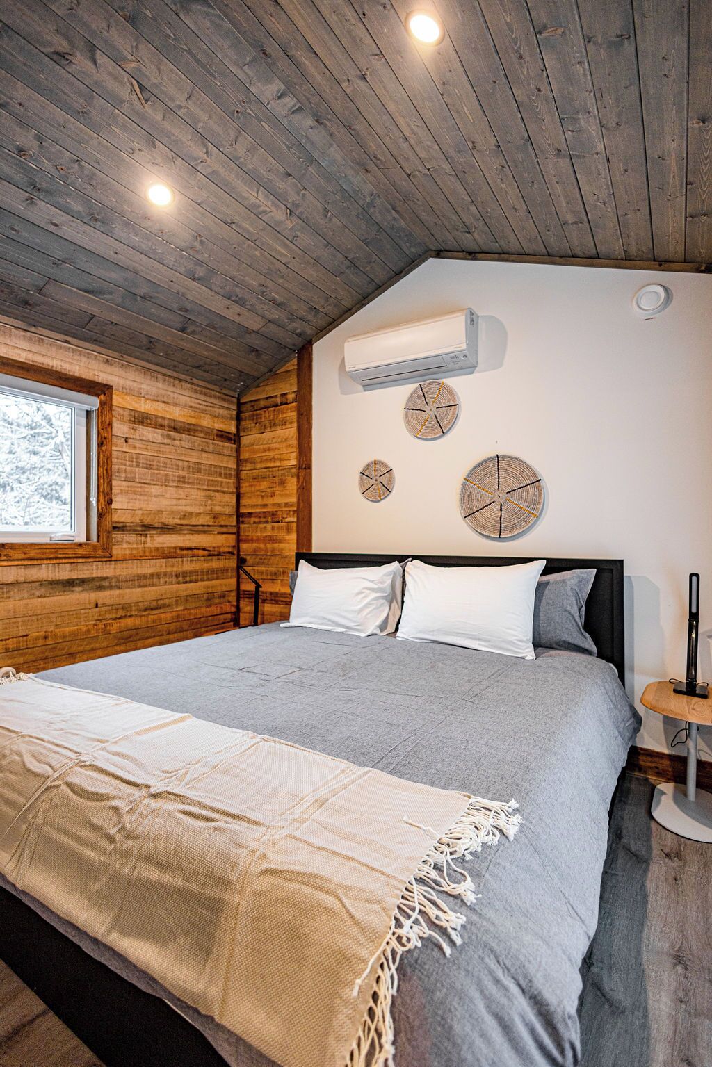 A bedroom with a wooden ceiling, a bed with grey bedding, a white air conditioner, and three wall-mounted clocks.