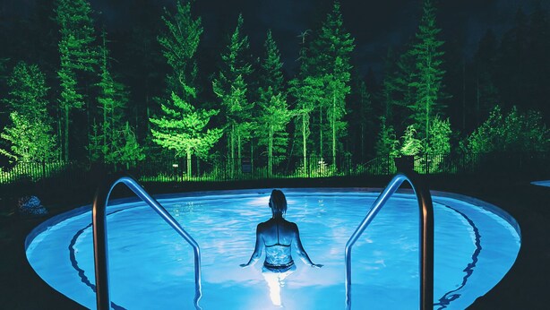 Une personne médite dans une piscine circulaire entourée de barrières métalliques, le tout étant situé au milieu d’une forêt illuminée par des lumières vertes.