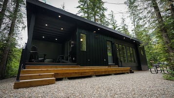 A modern cabin with a wooden deck and large windows, surrounded by trees.