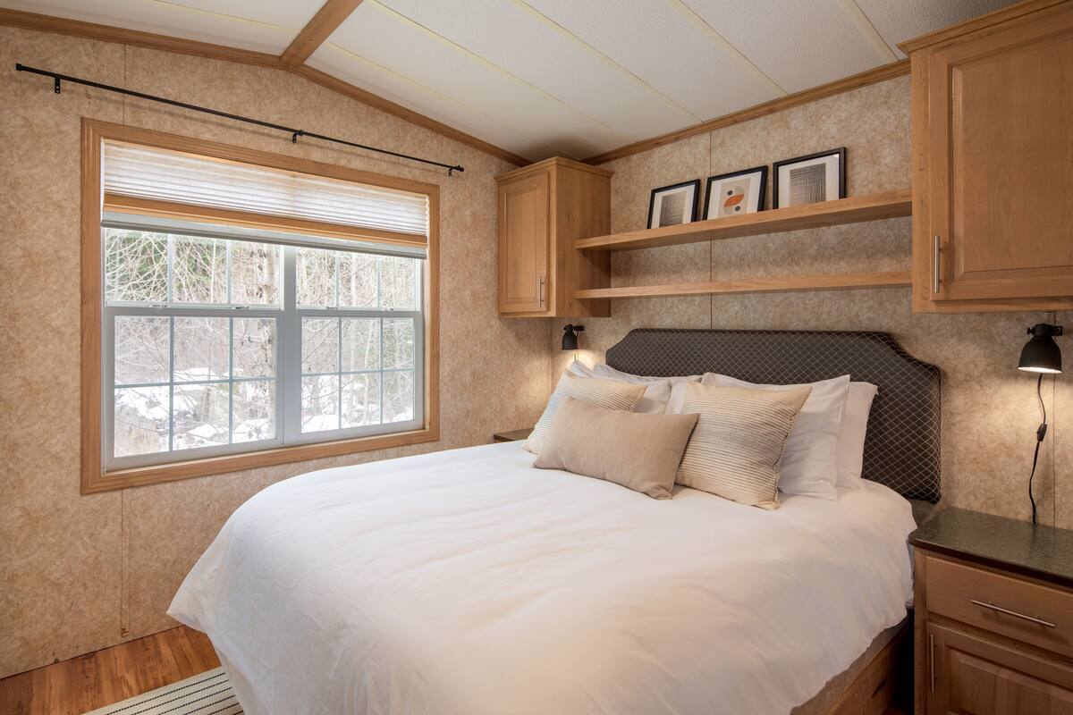 Un dormitorio con una cama grande, cabecera de madera, una ventana con persianas y estanterías empotradas con cuadros enmarcados.