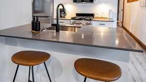 Une cuisine moderne avec des armoires blanches, un réfrigérateur en acier inoxydable, un four à micro-ondes et une cuisinière. On y trouve deux tabourets avec des assises marron et des pieds en métal noir.